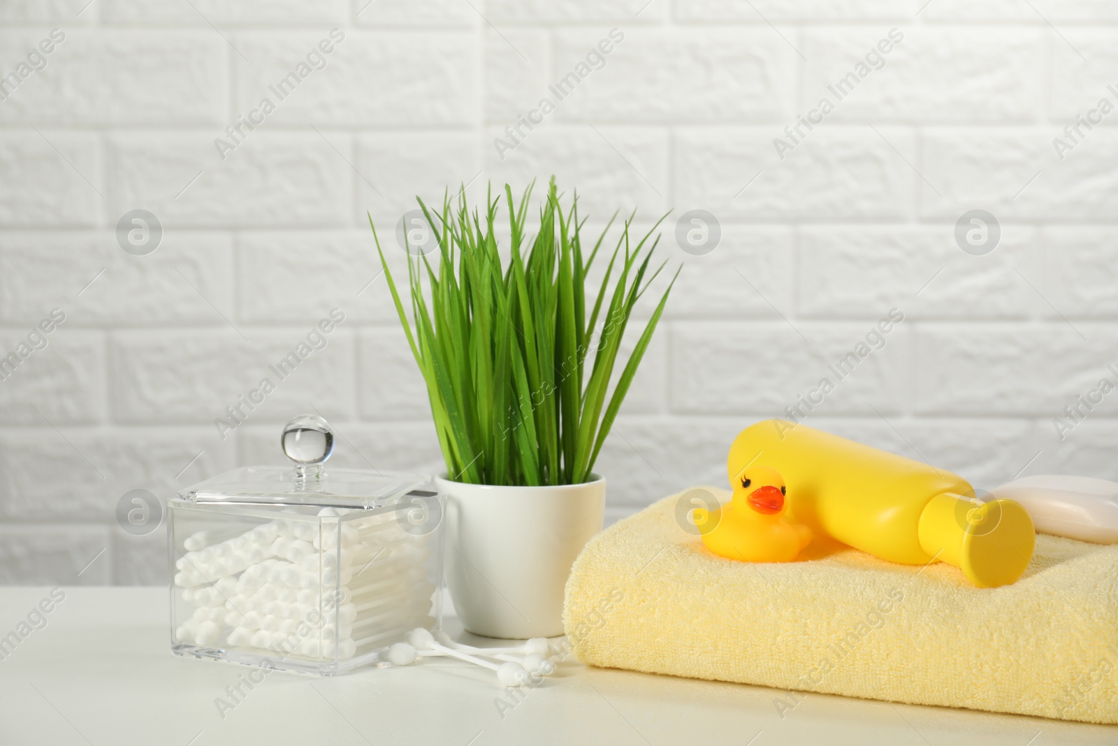 Photo of Baby bath accessories. Towel, cosmetic products, cotton swabs and toy duck on white table against brick wall