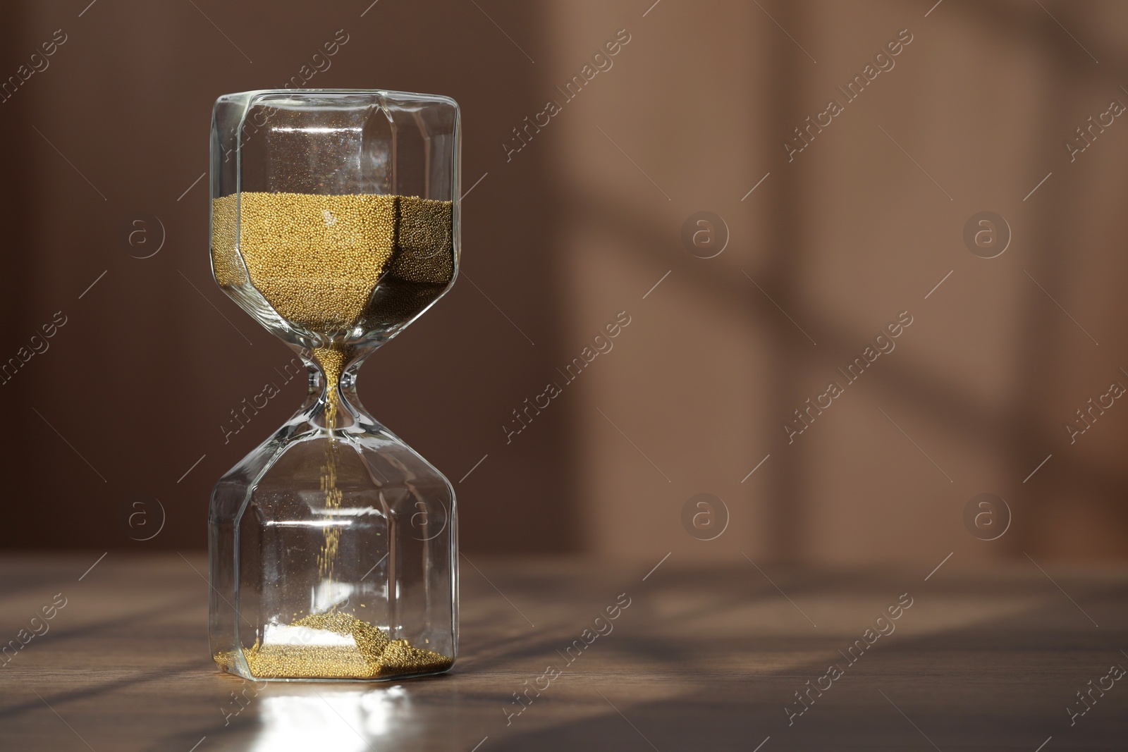 Photo of Hourglass with flowing gold sand on wooden table against brown background, space for text