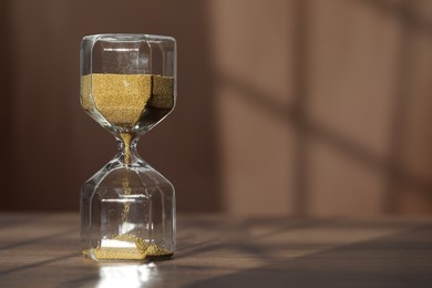 Hourglass with flowing gold sand on wooden table against brown background, space for text