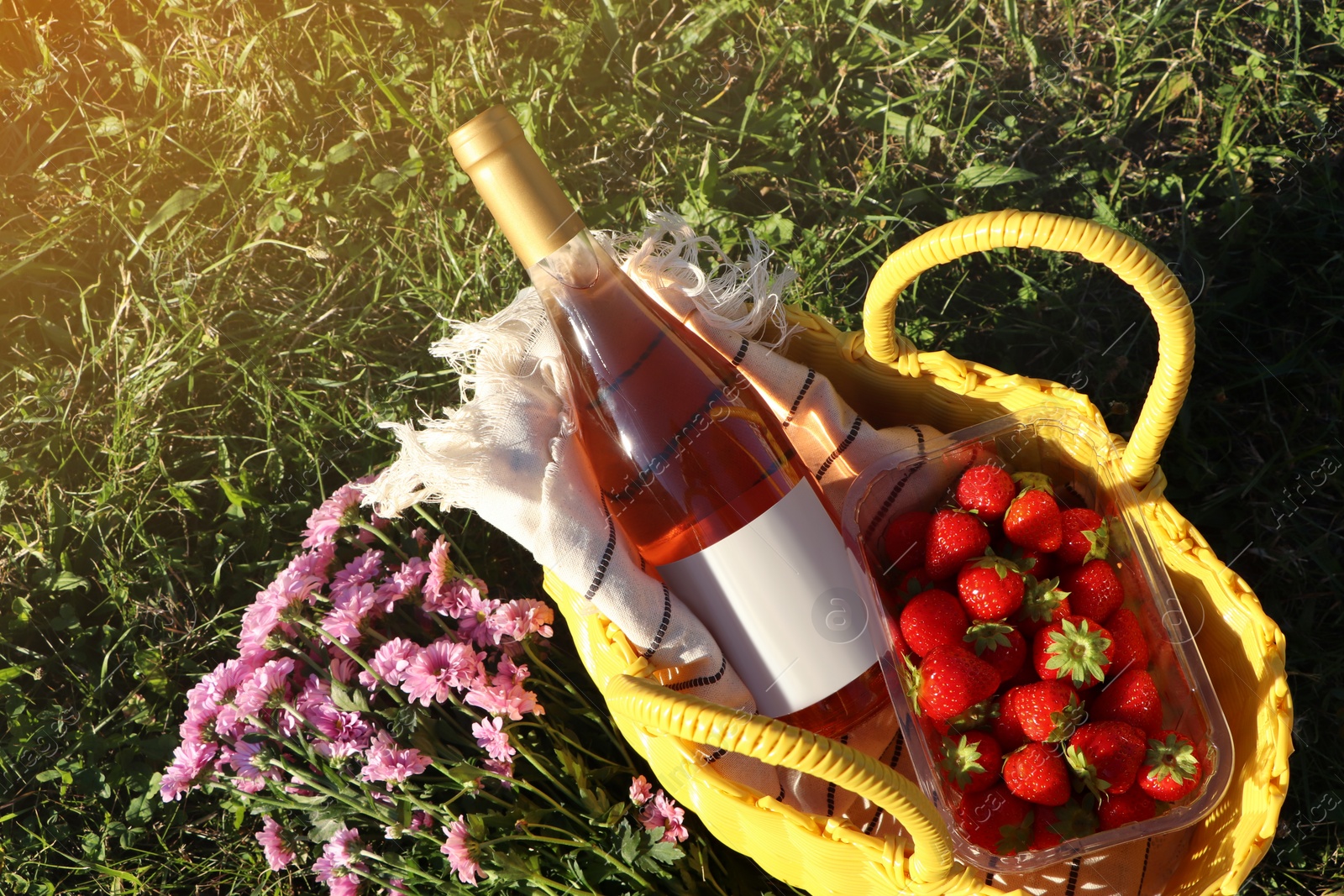 Photo of Yellow wicker bag with bottle of wine, strawberries, picnic blanket and beautiful flowers on green grass outdoors