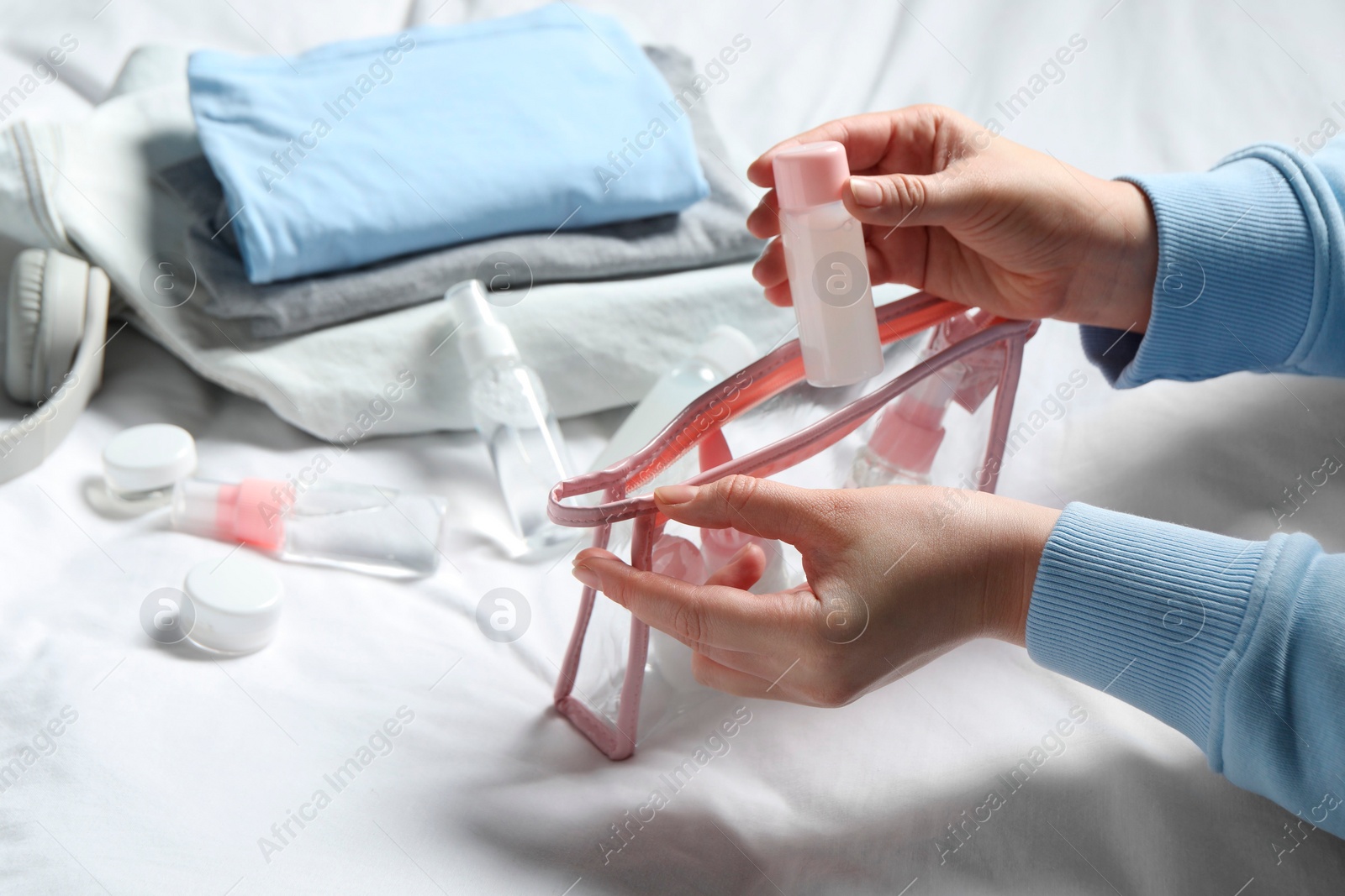 Photo of Cosmetic travel kit. Woman putting small bottle with personal care product into plastic bag on bed, closeup