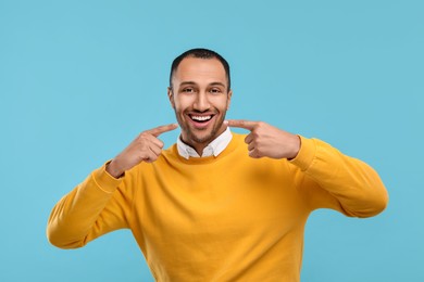 Smiling man pointing at his healthy clean teeth on light blue background