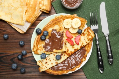 Photo of Tasty crepes with chocolate paste, banana and berries served on wooden table, flat lay