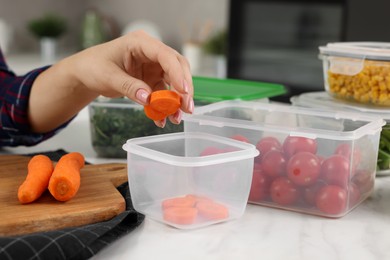 Woman putting fresh carrot into plastic container at white marble table, closeup. Food storage