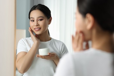 Photo of Happy woman applying face cream near mirror at home