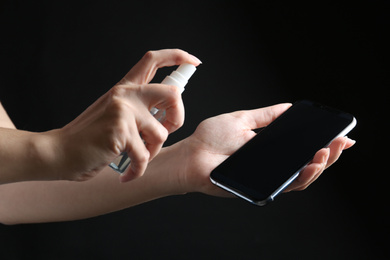 Photo of Woman spraying antiseptic onto smartphone against black background, closeup