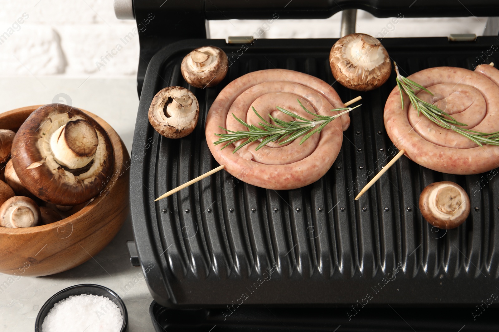 Photo of Electric grill with homemade sausages, rosemary and mushrooms on light table