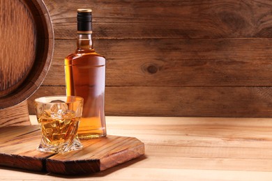 Photo of Whiskey with ice cubes in glass, bottle and barrel on wooden table, space for text