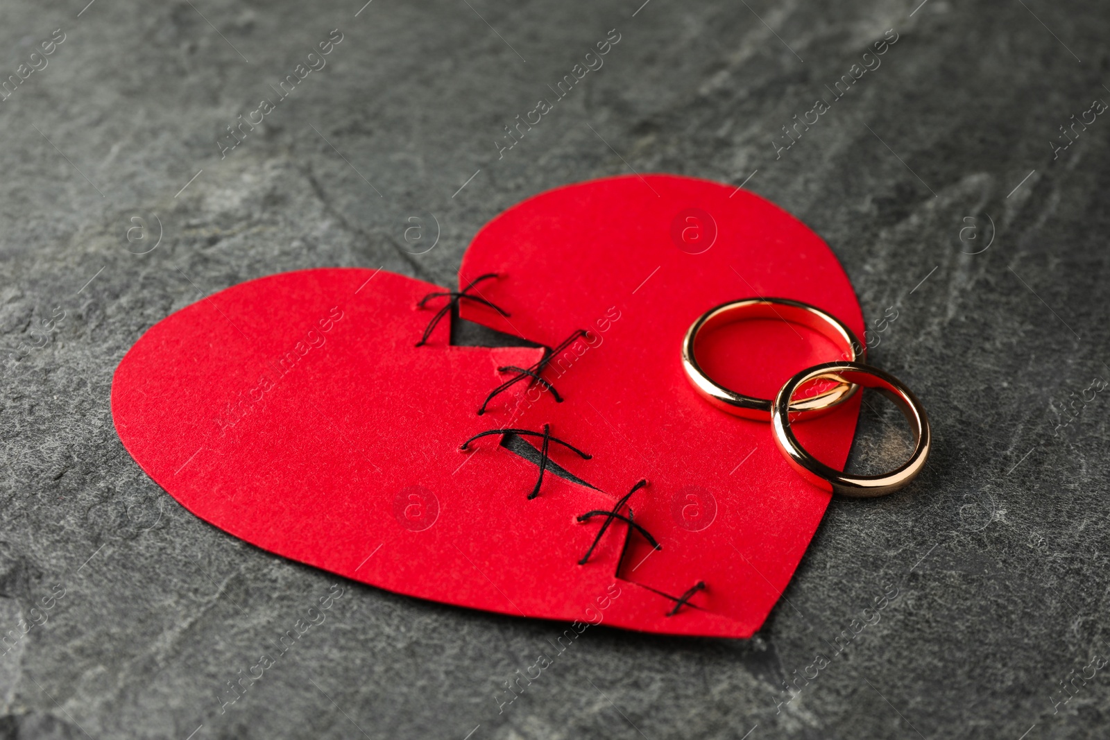 Photo of Broken heart. Torn red paper heart sewed with thread and wedding rings on grey table, closeup