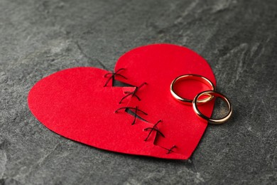 Broken heart. Torn red paper heart sewed with thread and wedding rings on grey table, closeup