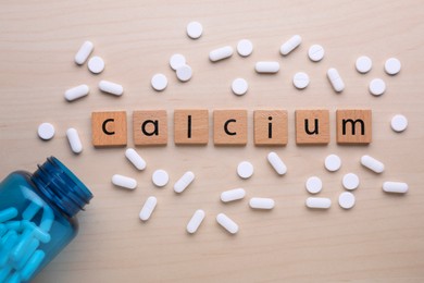 Photo of Word Calcium made of cubes with letters, medical bottle and pills on wooden table, flat lay