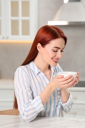 Photo of Happy woman with red dyed hair enjoying cup of drink at white table in kitchen
