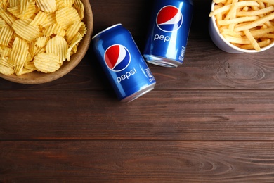 Photo of MYKOLAIV, UKRAINE - FEBRUARY 15, 2021: Cans of Pepsi and snacks on wooden table, flat lay. Space for text