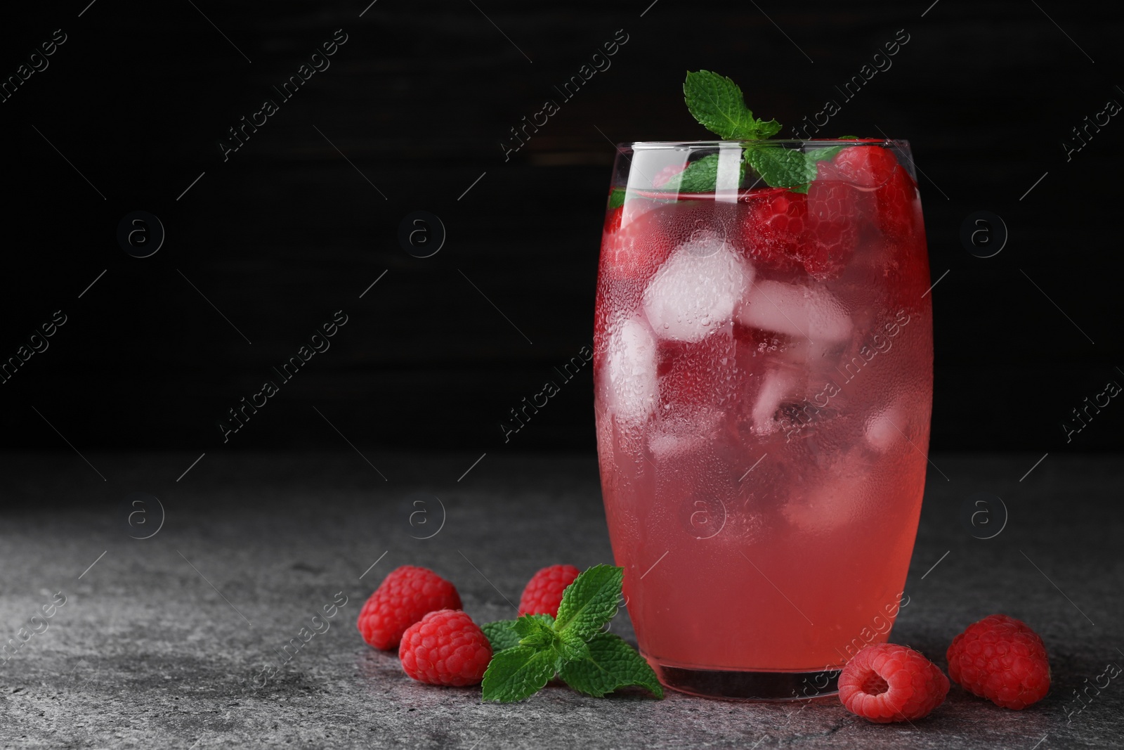 Photo of Glass of refreshing drink with raspberry and mint on grey stone table against dark background, space for text