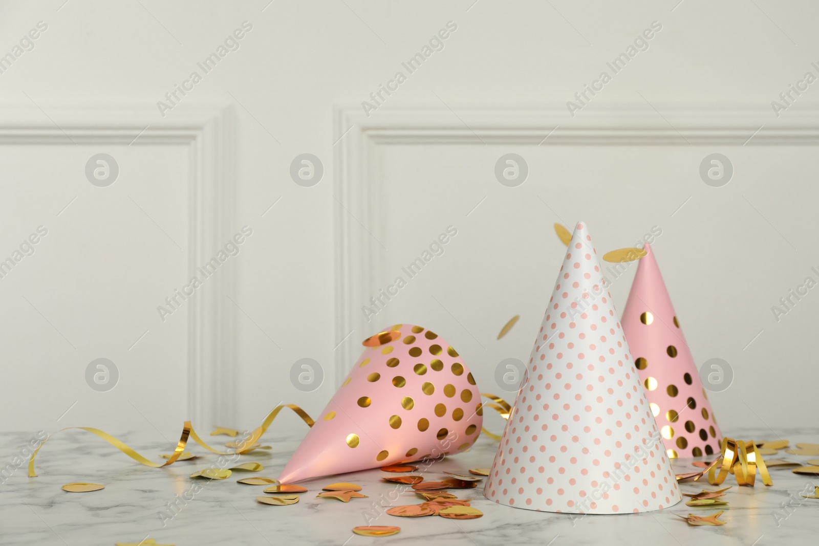 Photo of Shiny confetti falling on marble table with party hats near white wall indoors. Space for text