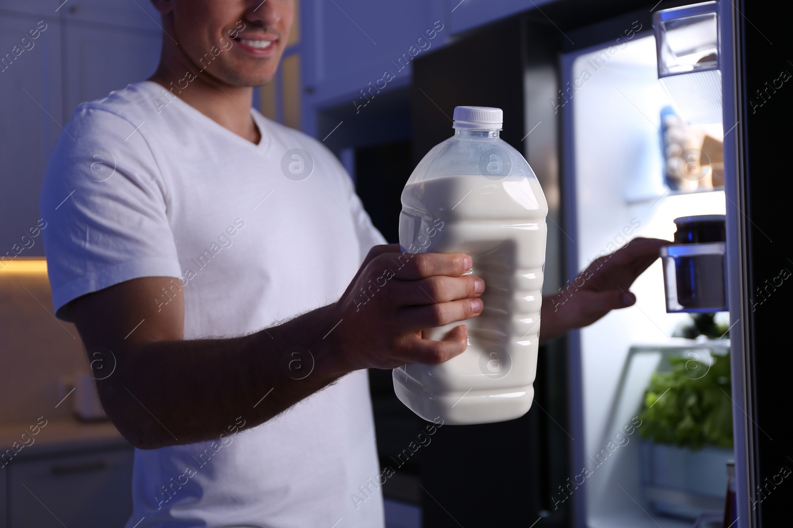 Photo of Man holding gallon bottle of milk near refrigerator in kitchen at night, closeup