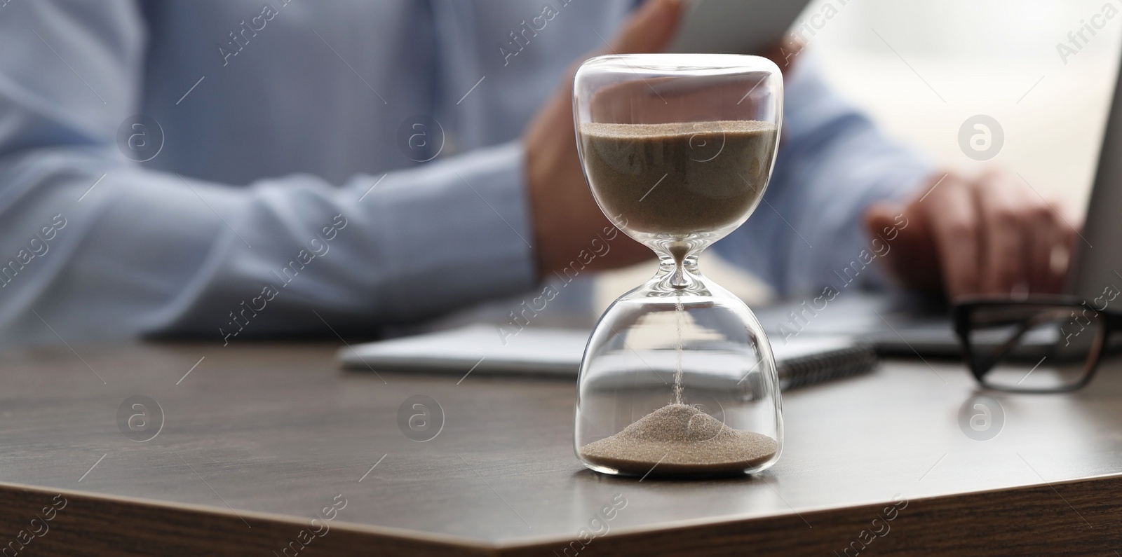 Photo of Hourglass with flowing sand on desk. Man using smartphone while working on laptop indoors, selective focus