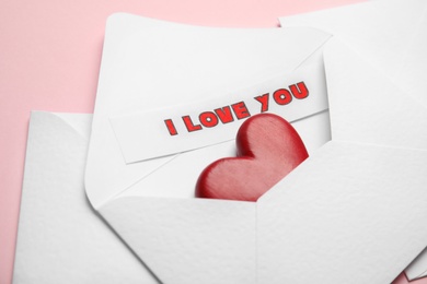Sheet of paper with phrase I Love You and decorative heart in envelope on pink background, closeup