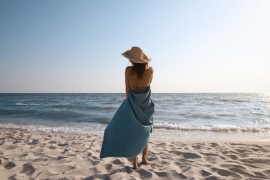 Woman with beach towel and straw hat on sand near sea, back view