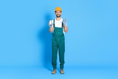 Professional repairman in uniform showing smartphone on light blue background