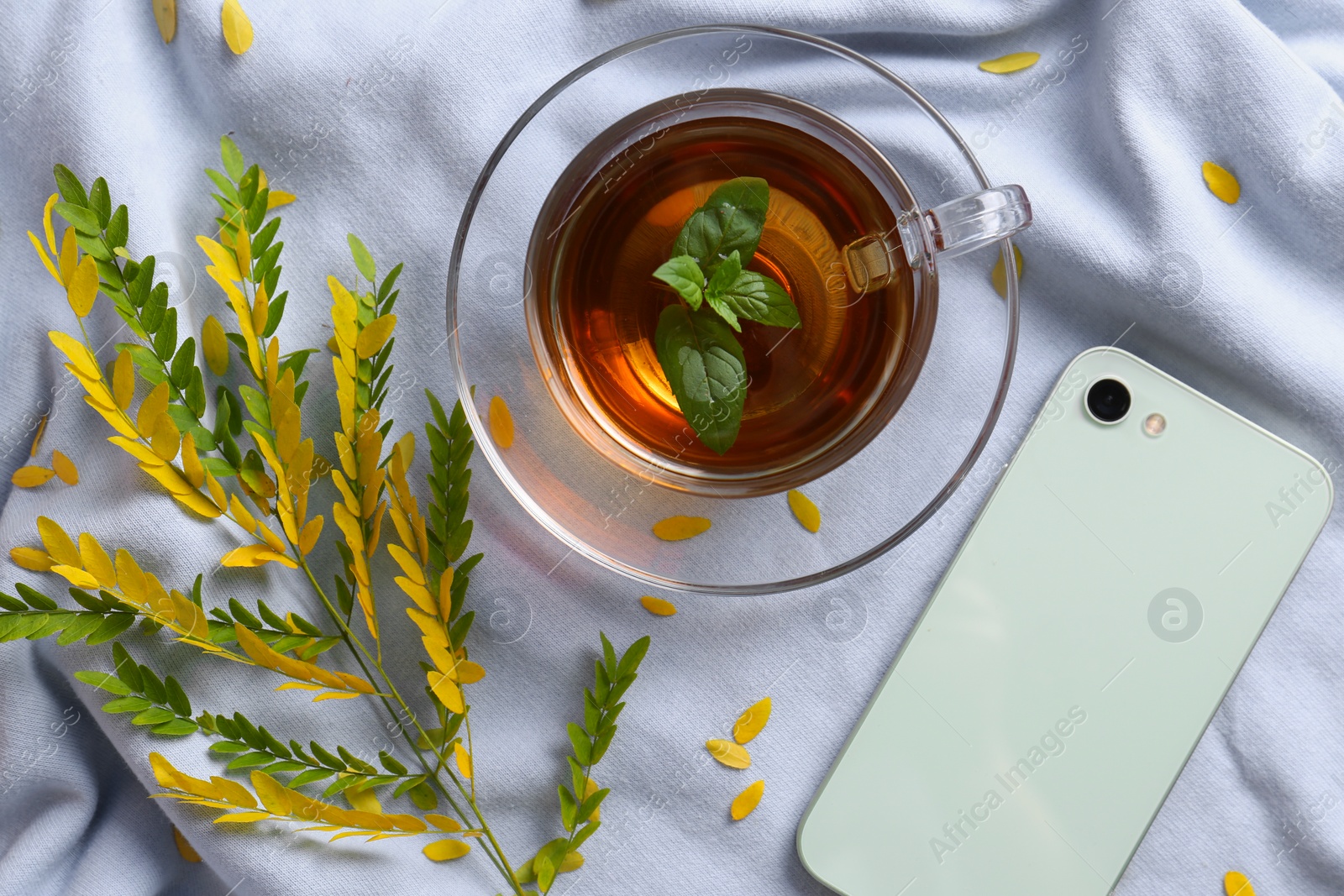 Photo of Cup of aromatic herb tea, smartphone and dry autumn leaves on white cloth, flat lay