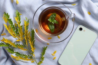 Cup of aromatic herb tea, smartphone and dry autumn leaves on white cloth, flat lay
