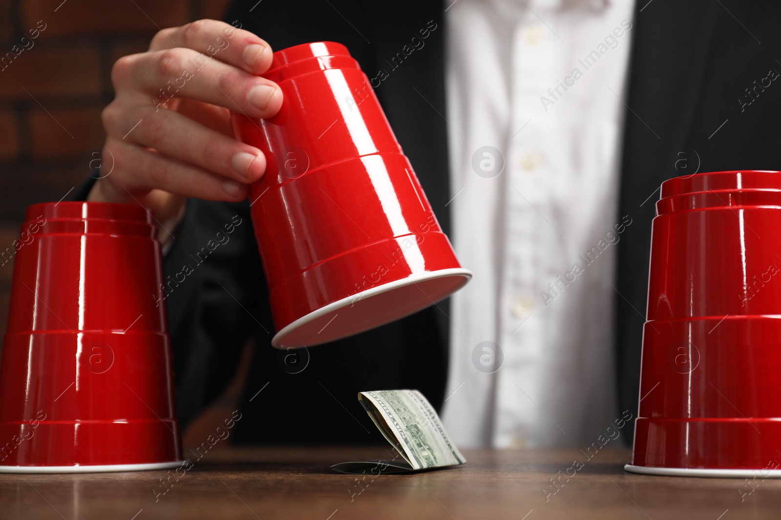 Photo of Shell game. Man showing dollar banknote under cup at wooden table, closeup