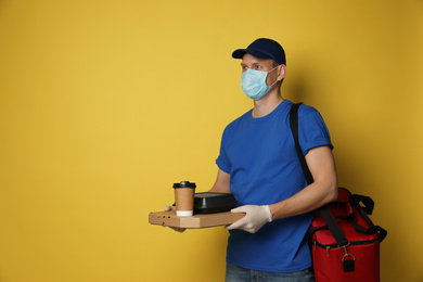 Courier in protective mask and gloves holding order on yellow background, space for text. Food delivery service during coronavirus quarantine