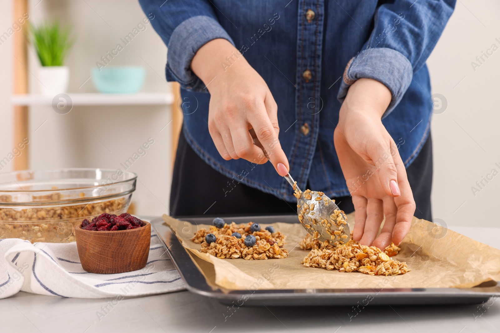 Photo of Making granola bars. Woman putting mixture of oat flakes, dry fruits and other ingredients onto baking tray at table in kitchen, closeup