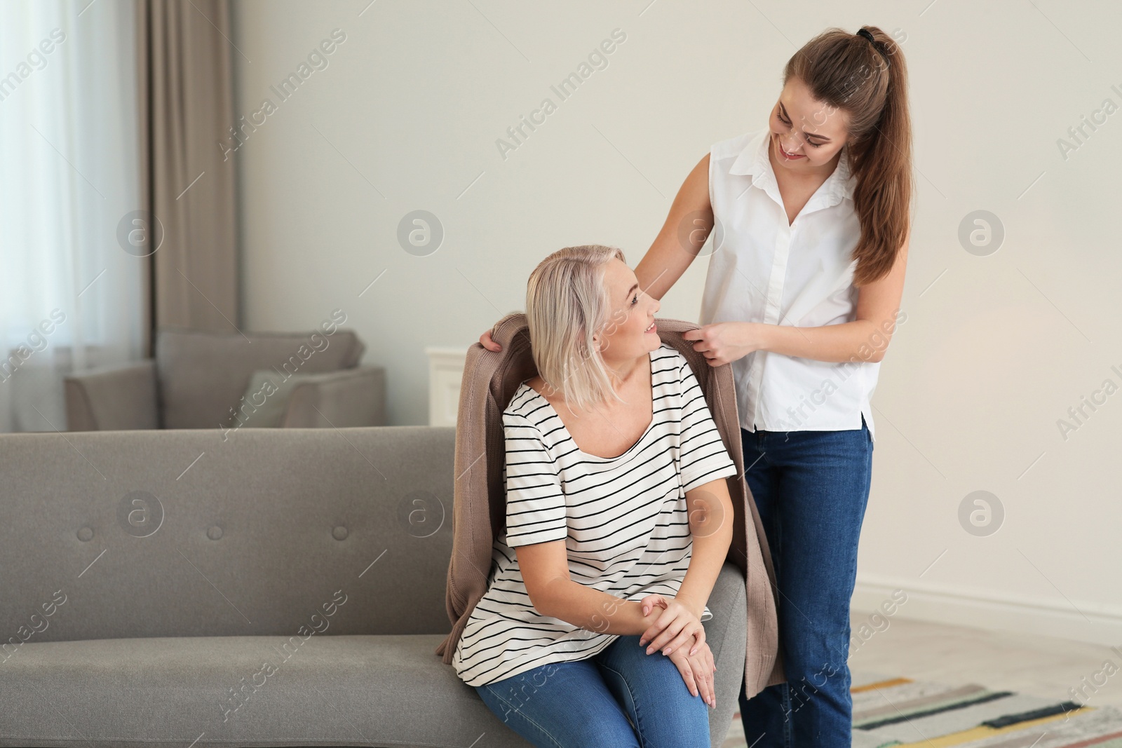 Photo of Young woman covering her mother with warm cardigan at home. Family help