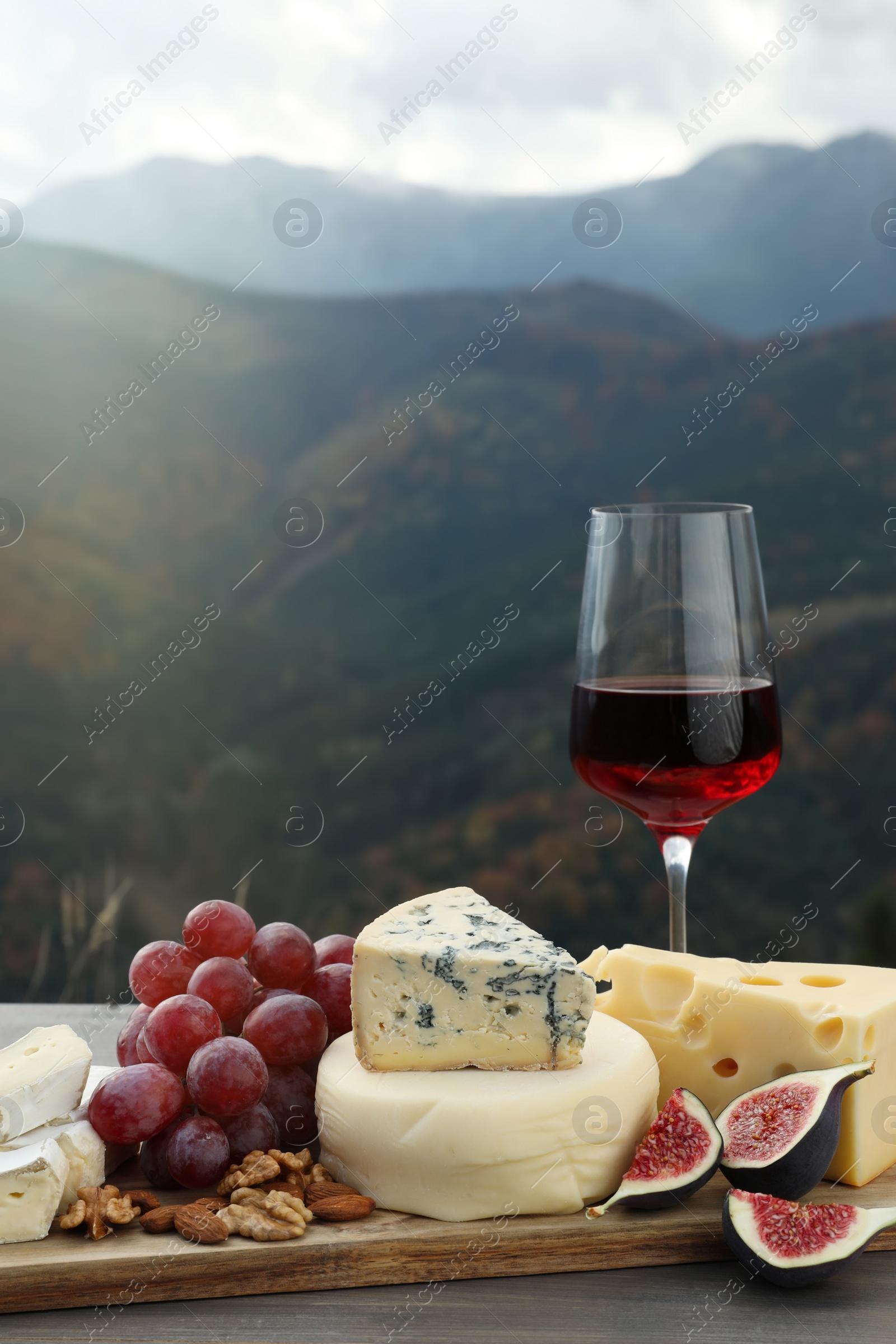 Photo of Different types of delicious cheeses, fruits and wine on wooden table against mountain landscape