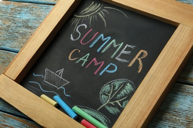 Photo of Blackboard with text SUMMER CAMP, drawings and chalk sticks on wooden background, closeup