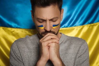 Sad man with clasped hands near Ukrainian flag, closeup