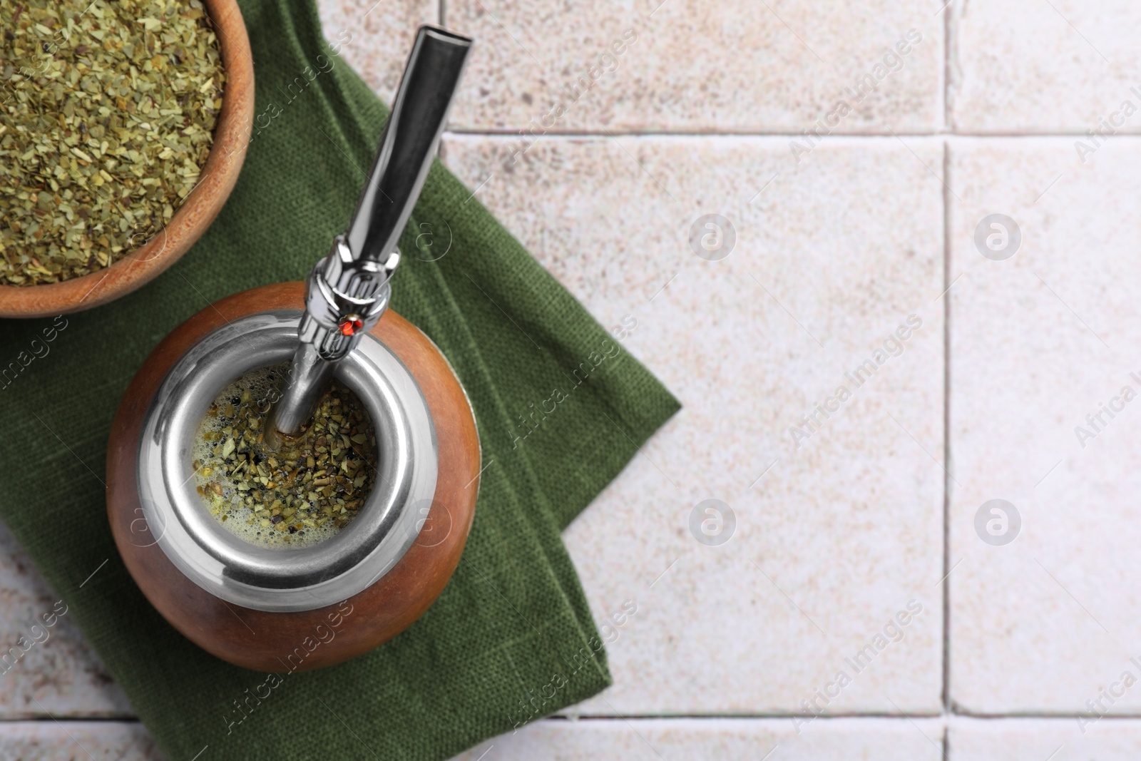 Photo of Calabash with bombilla, bowl of mate tea leaves and napkin on tiled table, flat lay. Space for text