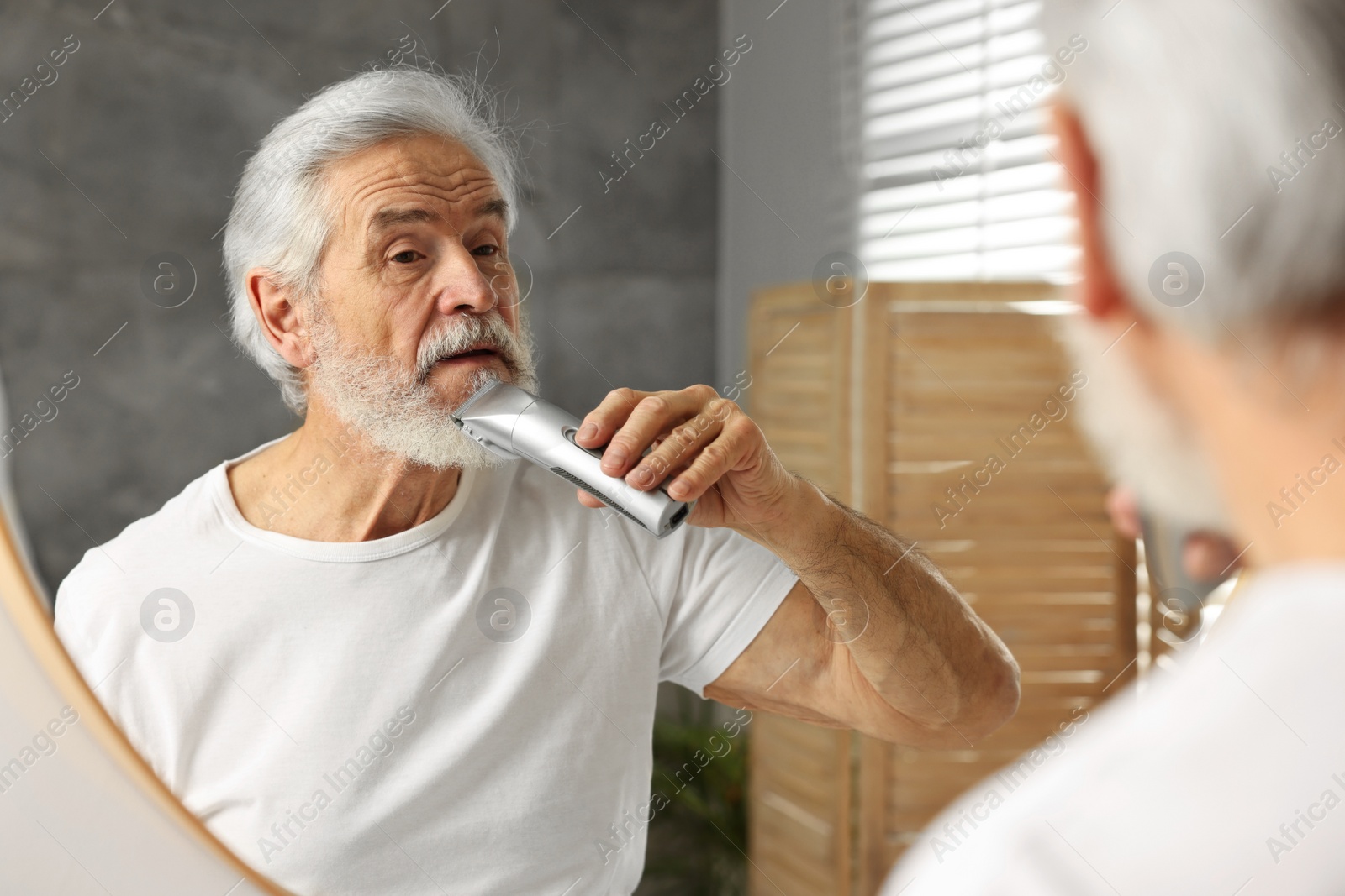 Photo of Senior man trimming beard near mirror in bathroom