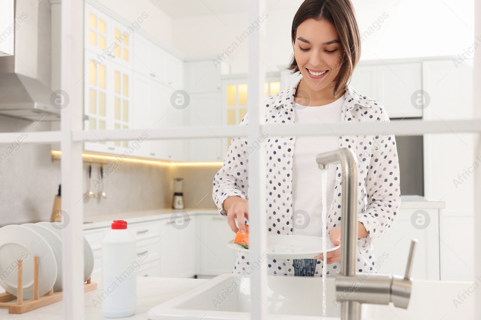 Photo of Happy young woman washing plate above sink in modern kitchen