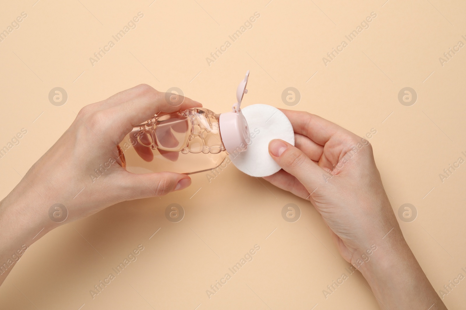 Photo of Woman pouring micellar water from bottle onto cotton pad against beige background, closeup