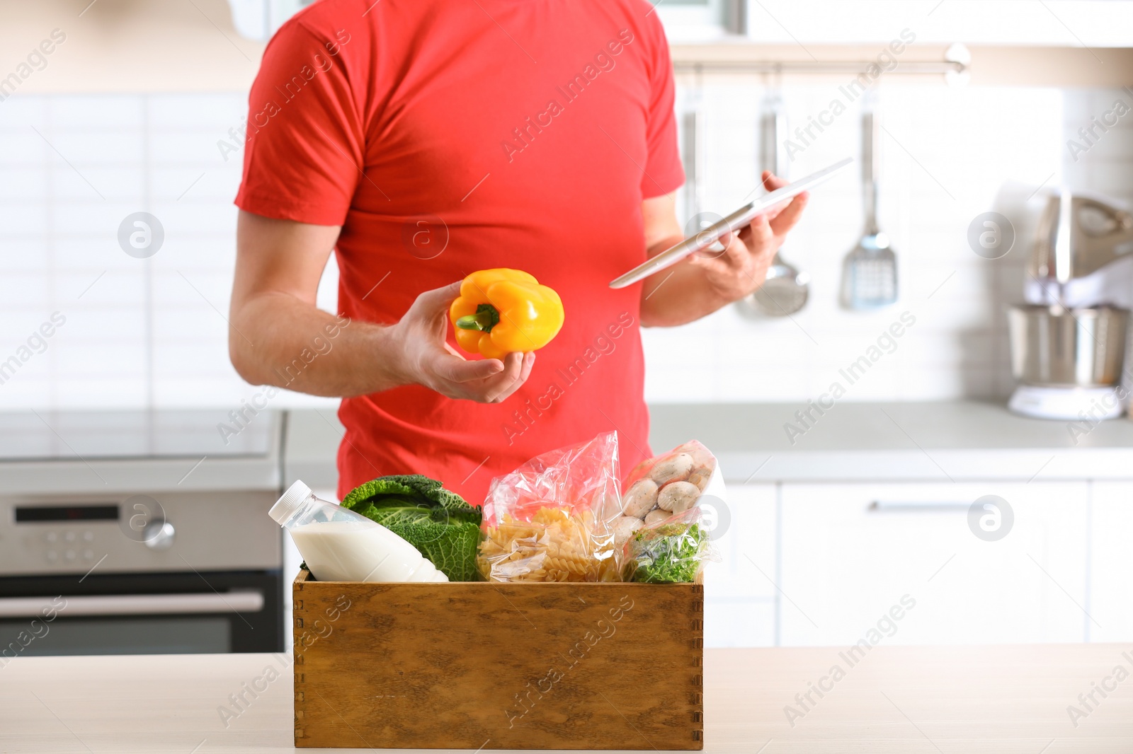 Photo of Man with wooden crate full of products and tablet in kitchen. Food delivery service