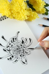 Photo of Woman drawing chrysanthemum in sketchbook at white table, closeup