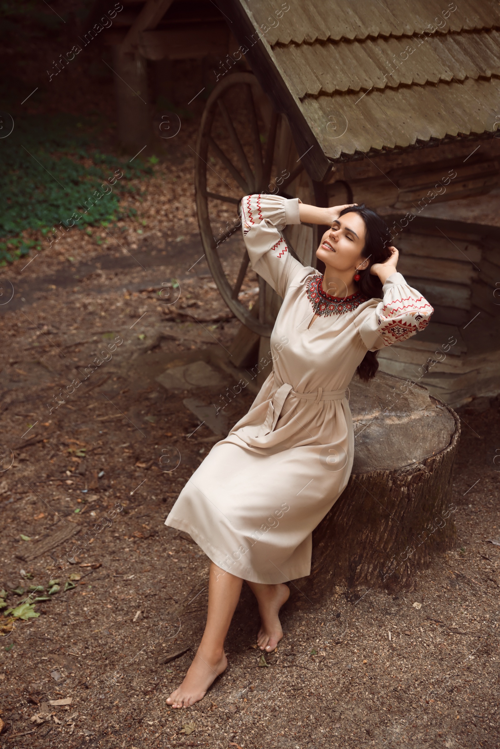 Photo of Beautiful woman wearing embroidered dress sitting near old wooden well in countryside. Ukrainian national clothes