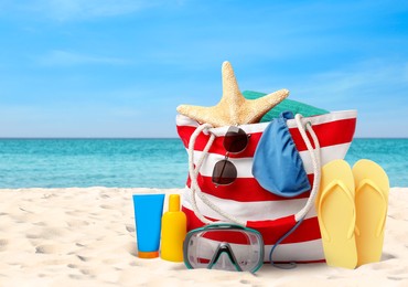 Image of Stylish bag with different accessories on sandy beach near ocean