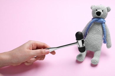 Photo of Woman pretending to test teddy bear's reflexes with hammer on pink background, closeup. Nervous system diagnostic