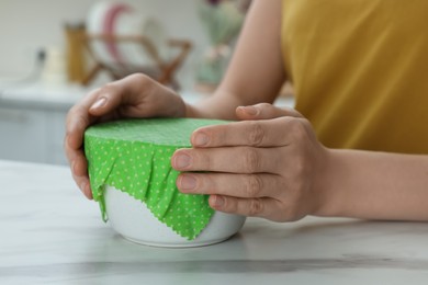 Photo of Woman holding bowl covered with beeswax food wrap at white marble table in kitchen, closeup