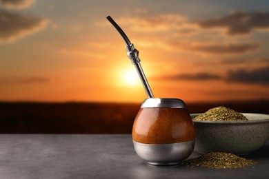 Image of Calabash with mate tea and bombilla on light grey table outdoors at sunset. Space for text