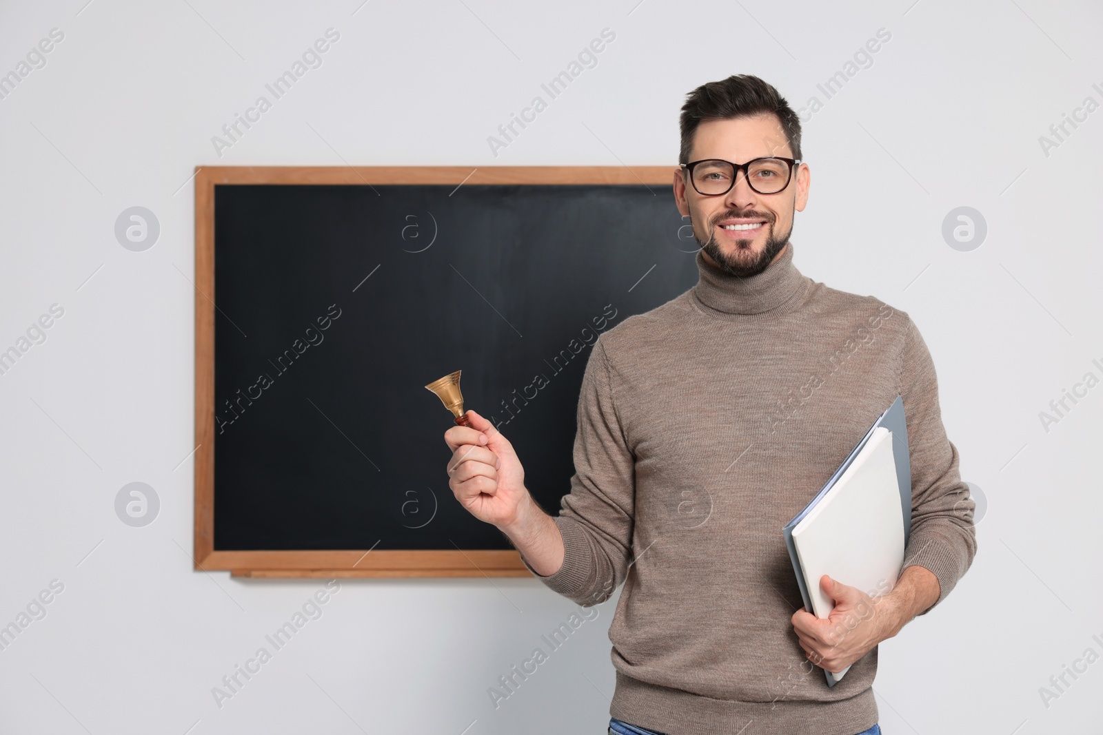 Photo of Teacher with school bell near black chalkboard indoors. Space for text