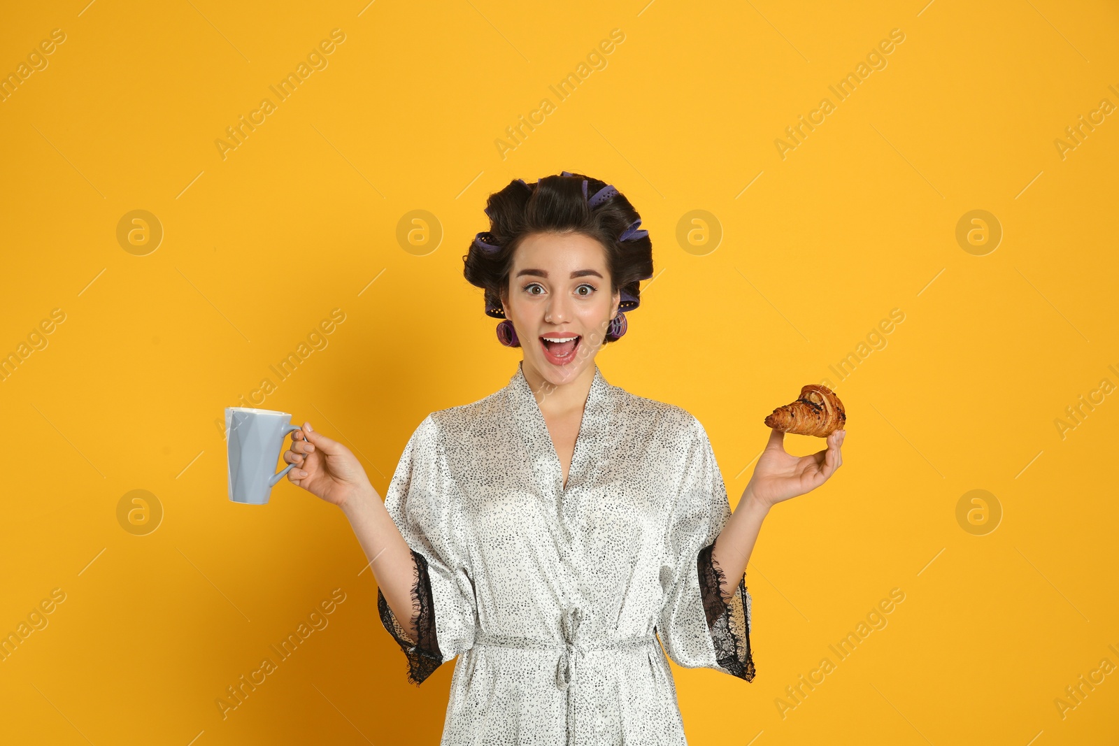 Photo of Excited young woman in silk bathrobe with hair curlers having breakfast on orange background