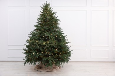 Photo of Beautiful Christmas tree decorated with festive lights near white wall indoors, space for text