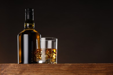 Whiskey with ice cubes in glass and bottle on wooden table, low angle view. Space for text