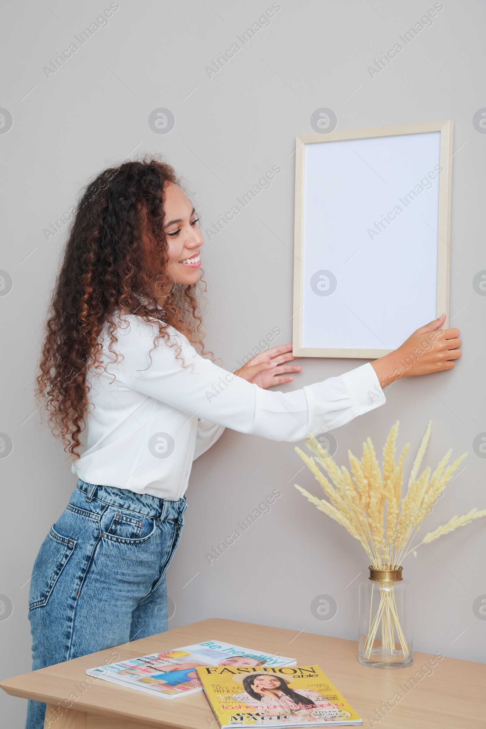 Photo of African American woman hanging empty frame on pale rose wall over table in room. Mockup for design
