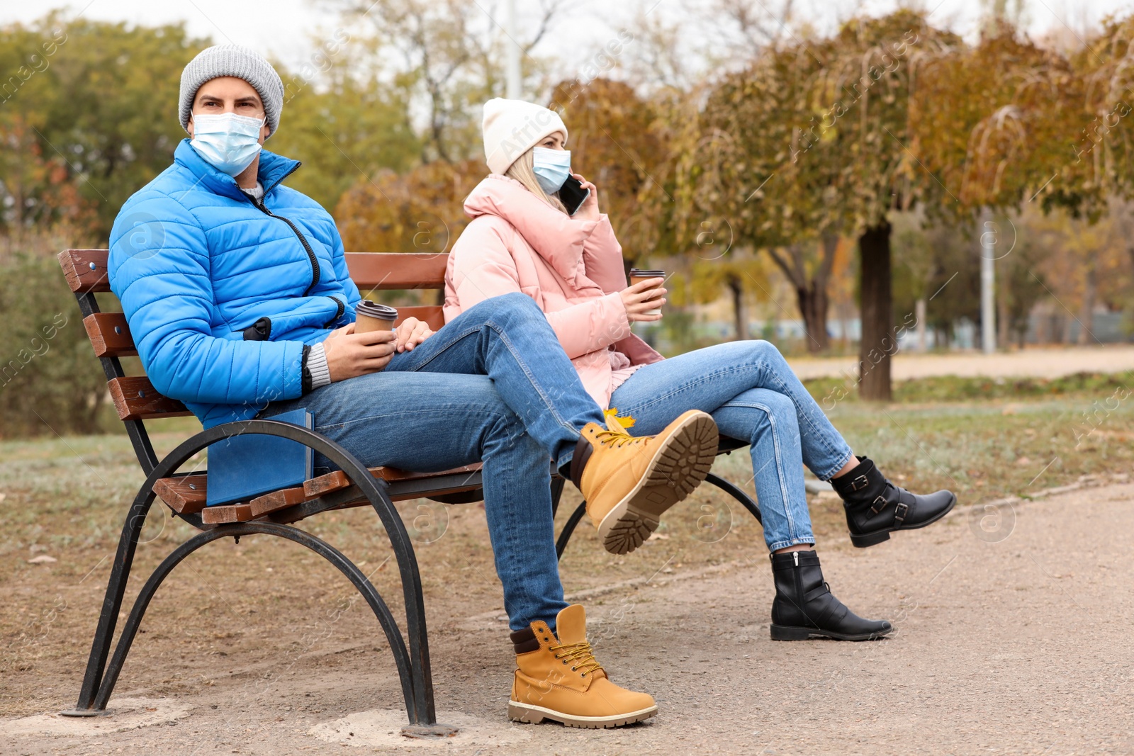 Photo of People in medical masks keeping distance while sitting on bench outdoors. Protective measures during coronavirus quarantine